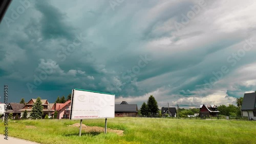 POV shot from a moving vehicle passing a quiet rural village with houses and green fields under a dramatic, stormy sky. The video captures a powerful contrast between peaceful countryside and