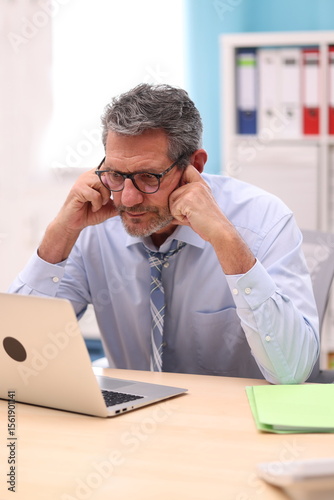 Office worker in shirt and tie sits at the desk, supports his head with both hands and looks concentratedly at his laptop