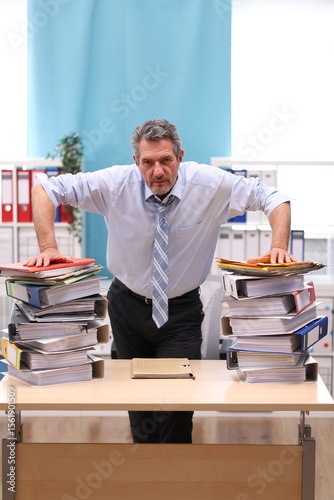Office worker in shirt and tie stands in the office in front of stacks of files and looks very annoyed and exhausted