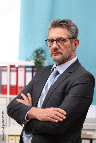 Office worker with glasses and suit has his arms crossed in front of his body and looks sternly and dominantly to the side