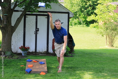 Middle aged man in a blue shirt is throwing corn hole bags during a backyard game