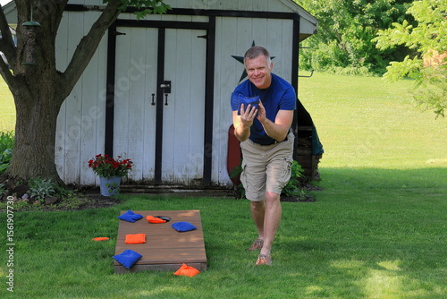 Middle aged man in a blue shirt is throwing corn hole bags during a backyard game