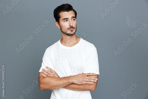 Thoughtful young caucasian man with stylish haircut wears white t-shirt standing against gray background, arms crossed, showcasing a contemplative expression.