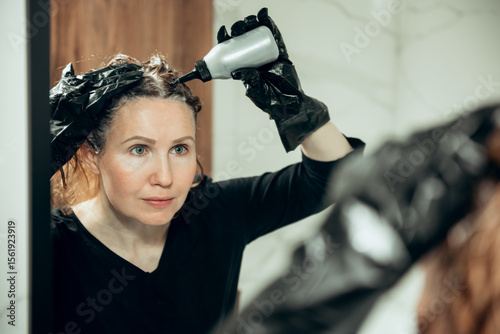 Middle-aged woman dyes her hair on her own in the bathroom.