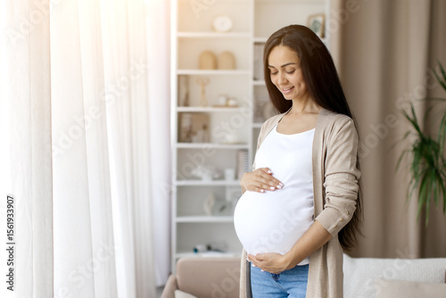 Картината върху платно Pregnant woman embracing belly and smiling peacefully while standing at home near window, cradling her belly
