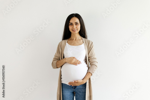 Εκτύπωση καμβά Young pregnant woman smiling and gently holding her belly while standing at home
