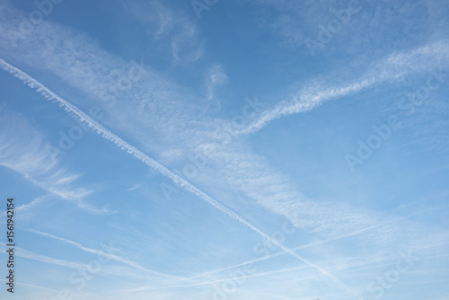 Fototapeta Naklejka Na Ścianę i Meble -  A clear blue sky with soft clouds and distinct plane tracks streaking across, creating a minimalist yet dynamic composition. A serene and airy view of the open sky.