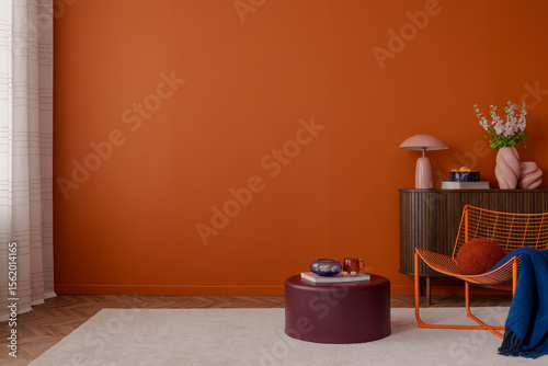 Minimalist and colorful interior with a vivid orange wall, dark wooden cabinet, and sculptural decor pieces. A burgundy ottoman and orange wire chair add a playful modern touch. Copy space.