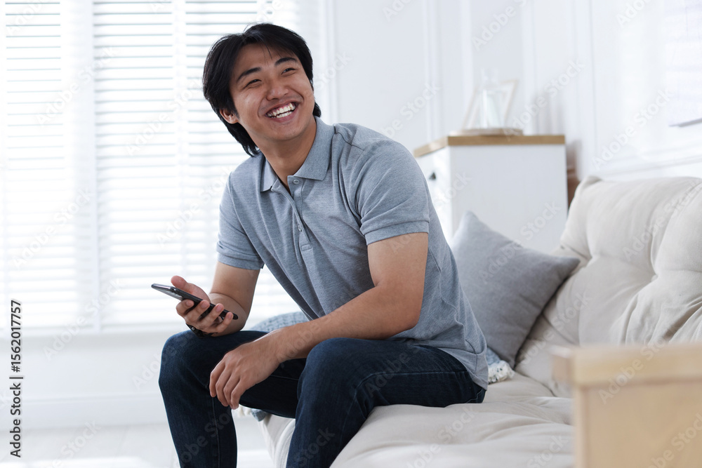 Obraz premium Smiling Asian man with smartphone on sofa at home