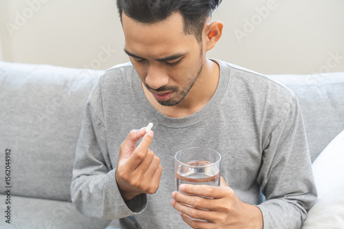 Sick asian young man, male holding tablet pill on hand, painkiller medicine from fever, stomach pain, headache for treatment, take drug or vitamin with glass of water on at home. Health care concept.