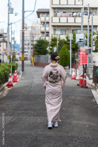 A woman wearing a traditional pink kimono with a floral obi walks down a quiet urban street in Japan. Rear view.