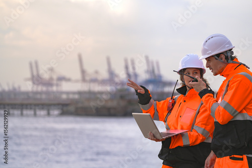 Cuadro en lienzo Two engineers wearing safety uniforms and hard hats communicate using a radio while reviewing data on a laptop by the sea during sunset