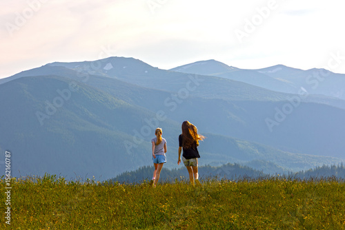 Two Girls Running Freely in Mountain Meadow. Back view of two girls joyfully running across a grassy hilltop with vast forested mountain ranges and soft sunlight in the background.