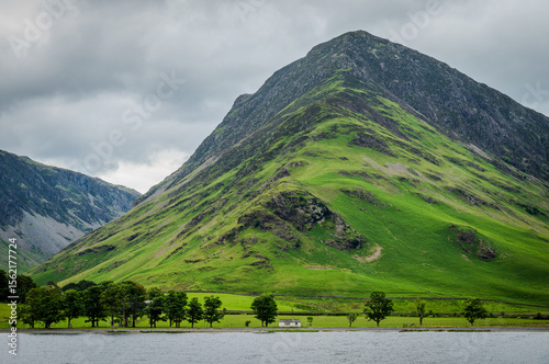 A moody summer landscape featuring a towering fell with a small cabin nestled at its base, captured on a cloudy day at Buttermere lake