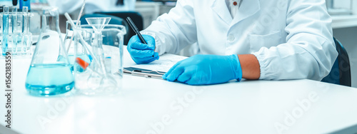 Young Asian male scientist recording data in a modern laboratory. Wearing safety goggles and gloves, surrounded by laboratory glassware. Biotechnology research and scientific documentation in progress photo