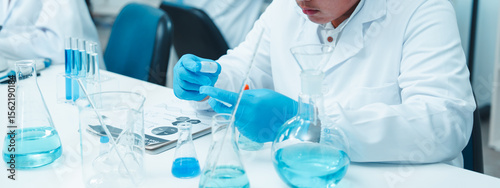 Asian male scientist wearing safety goggles and gloves working with blue liquid in a laboratory. Other researchers are working in the background in a clean, modern scientific environment.