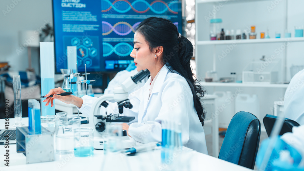 Fototapeta premium Asian female scientist in a white lab coat taking notes during a scientific experiment in a modern laboratory. Test tubes, microscope, and digital equipment are placed on the workbench.