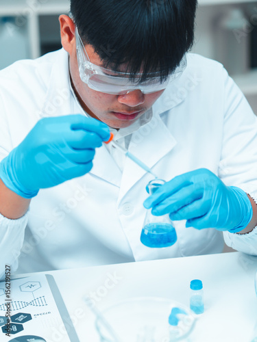Asian male scientist wearing lab coat and safety goggles using pipette to transfer blue liquid in modern laboratory. Beakers and flasks with chemical solutions are on the table.