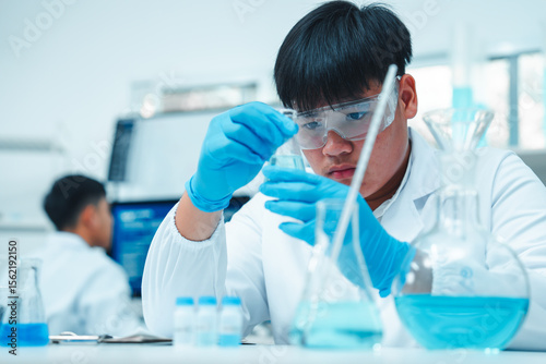 Asian male scientist wearing lab coat and safety goggles using pipette to transfer blue liquid in modern laboratory. Beakers and flasks with chemical solutions are on the table.