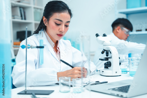 Asian female scientist in white lab coat taking notes beside a laptop in a clean laboratory. Laboratory glassware and scientific tools are on the table. Focused and professional atmosphere.