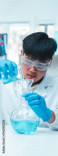 Male scientist in white lab coat and protective eyewear carefully pouring blue liquid into a glass flask using a funnel in a modern laboratory. Science experiment and chemical research in progress.
