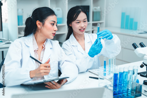 Two female scientists in lab coats working together in a science laboratory. One takes notes on a clipboard while the other examines a test tube. Collaborative STEM learning and research environment.