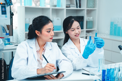 Two female scientists in lab coats working together in a science laboratory. One takes notes on a clipboard while the other examines a test tube. Collaborative STEM learning and research environment.