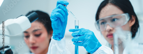 Two female scientists working in a modern laboratory. One is carefully transferring liquid with a pipette while the other observes and takes notes. STEM education and teamwork in a research setting.
