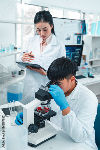Male scientist examining a specimen under a microscope while a female scientist records observations on a clipboard. Laboratory setting focused on scientific analysis, research, and teamwork.