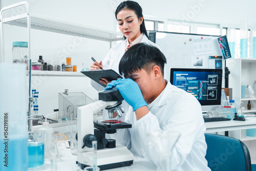 Male scientist examining a specimen under a microscope while a female scientist records observations on a clipboard. Laboratory setting focused on scientific analysis, research, and teamwork.