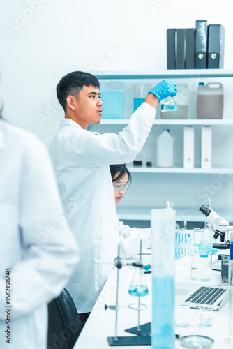 Male scientist holding a flask with blue liquid for examination in a laboratory. Other researchers work in the background. Scene reflects scientific research, analysis, and lab teamwork.