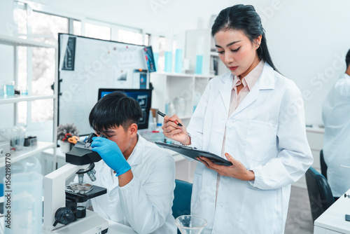 Male scientist examining a specimen under a microscope while a female scientist records observations on a clipboard. Laboratory setting focused on scientific analysis, research, and teamwork.