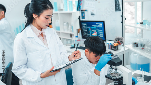 Scientist observes a sample under a microscope while a female colleague records data on a clipboard in a modern laboratory setting. Concept of scientific research, teamwork, and data analysis.