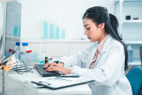 Female scientist in lab coat typing on a keyboard while analyzing research data on a computer. Laboratory equipment and chemical samples are visible, showing a scientific and focused work environment.