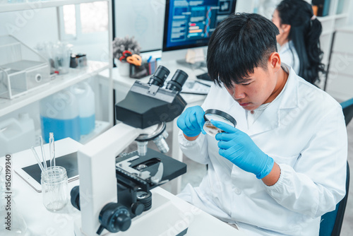 Young male scientist in lab coat examining a petri dish with a magnifying glass in a laboratory. Wearing blue gloves, focused on biological research. Microscope and lab tools visible on the desk.