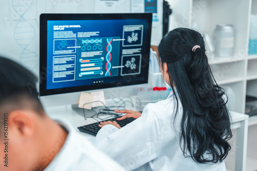 Female scientist in lab coat typing on a keyboard while analyzing research data on a computer. Laboratory equipment and chemical samples are visible, showing a scientific and focused work environment.