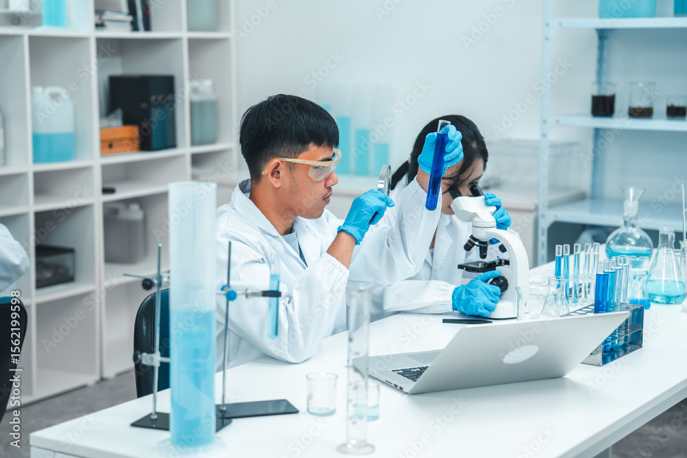 Fototapeta premium Two scientists in a laboratory working with test tubes. holds up a blue liquid sample while the woman observes attentively. Both wear lab coats, gloves, and safety goggles. Microscope in foreground.