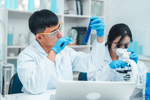 Two scientists in a laboratory working with test tubes. holds up a blue liquid sample while the woman observes attentively. Both wear lab coats, gloves, and safety goggles. Microscope in foreground.
