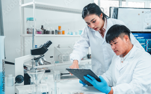 Female senior scientist supervising young male researcher in a modern lab. She reviews data on a clipboard while he listens attentively. Professional teamwork in scientific research setting.