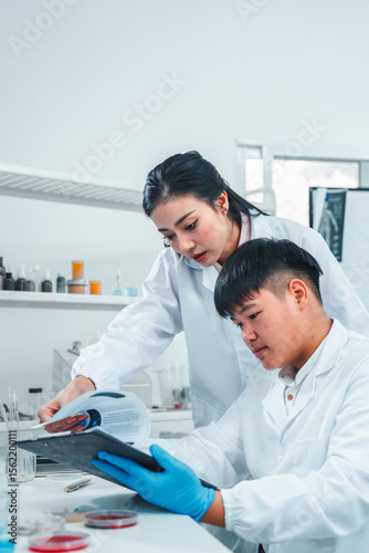 Female senior scientist supervising young male researcher in a modern lab. She reviews data on a clipboard while he listens attentively. Professional teamwork in scientific research setting.