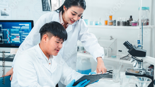 Female senior scientist supervising young male researcher in a modern lab. She reviews data on a clipboard while he listens attentively. Professional teamwork in scientific research setting.