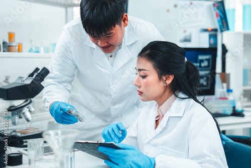 Young researchers collaborate on mold sample analysis in a science lab. The male scientist holds a petri dish while the female supervisor examines data. Team discovery and scientific breakthrough.