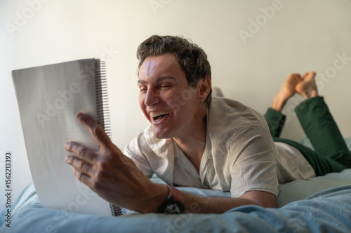 Mature man laughing while reading a notebook lying on bed
