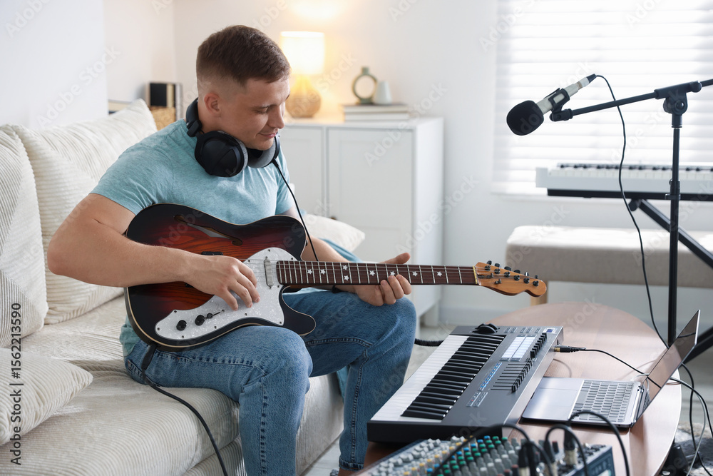 Obraz premium Man working with guitar and MIDI keyboard on sofa in home studio