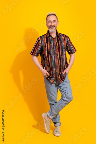 Charismatic fashionable gentleman in colorful shirt and jeans standing on bright yellow vertical background expressing cheerfulness