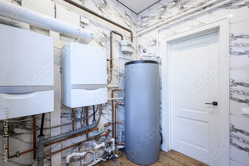 utility room with water heaters, copper piping, and a grey storage tank. The walls are tiled in a marble pattern, and a white door is present