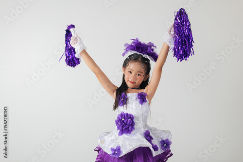 Little cheerleader girl in purple uniform with pom-poms, smiling joyfully. White studio background highlights her enthusiasm and team spirit. cheerleader kid
