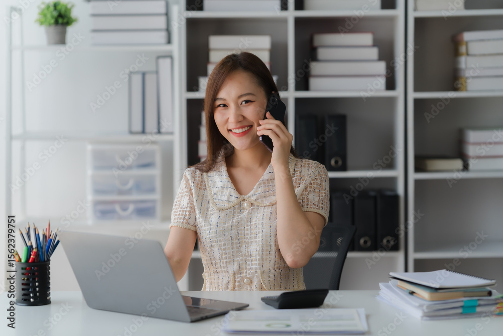 Obraz premium Asian Professional on Call: Radiant professional woman, engrossed in a mobile call at office desk. The scene exudes confidence, showcasing modern productivity amidst the company.