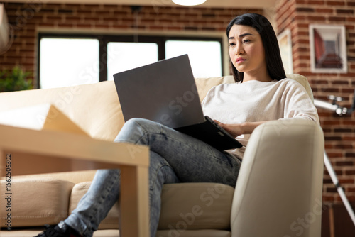 Serious female manager working remotely in brick wall apartment, typing on laptop and preparing business reports. Self employed asian woman showing productivity at home, reviewing company proposals