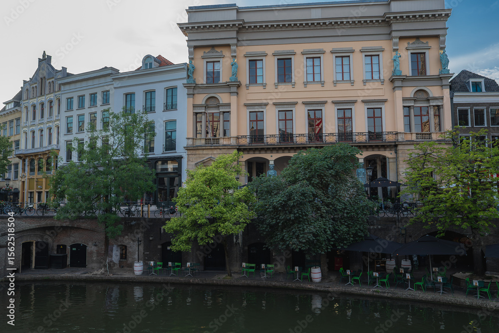 Obraz premium A canal in Utrecht, Netherlands, bordered by trees and historic buildings. Features a yellow building with statues and a walkway with outdoor seating.